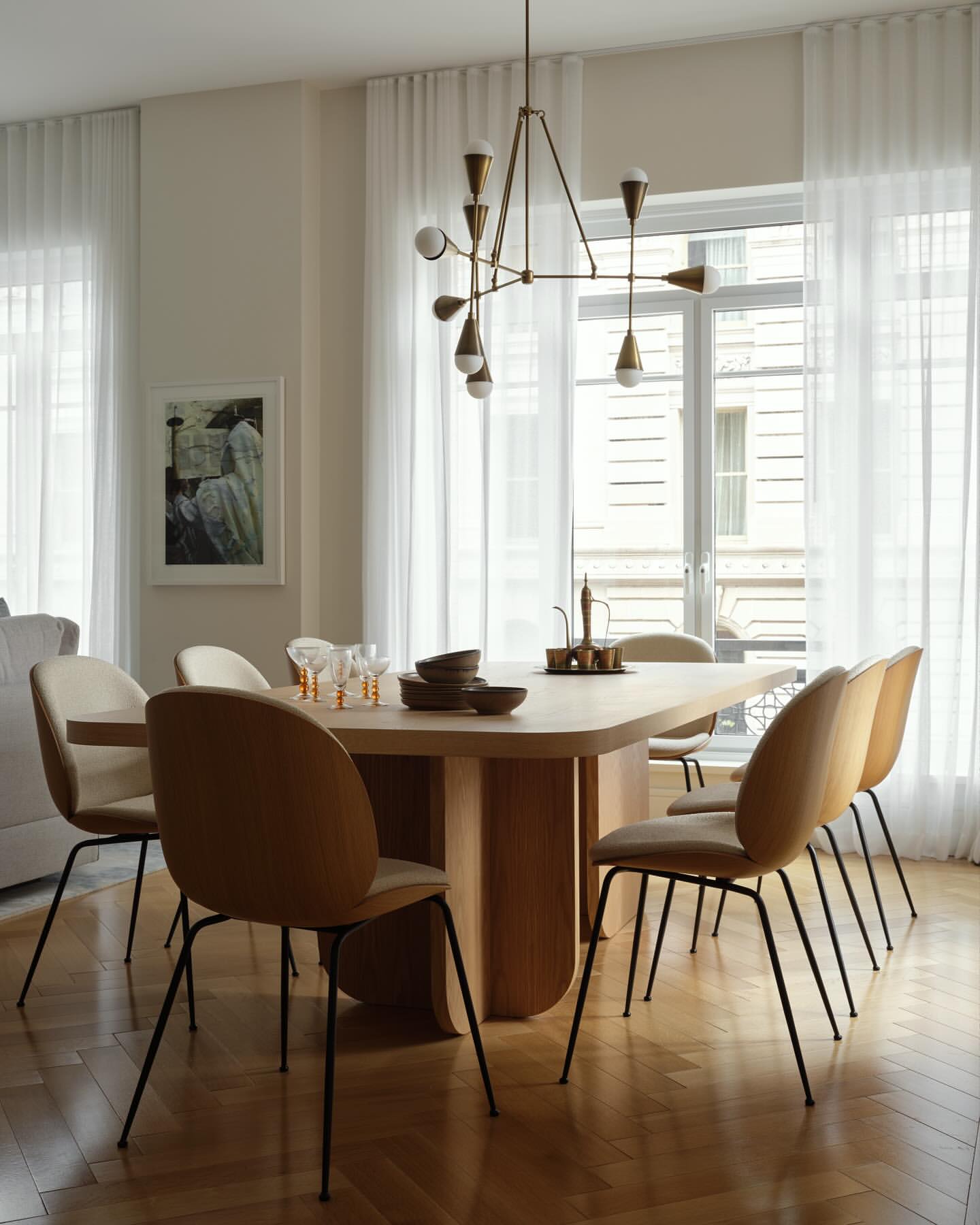 Modern dining room featuring a light wood table surrounded by tan chairs, complemented by a brass chandelier and sheer white curtains.