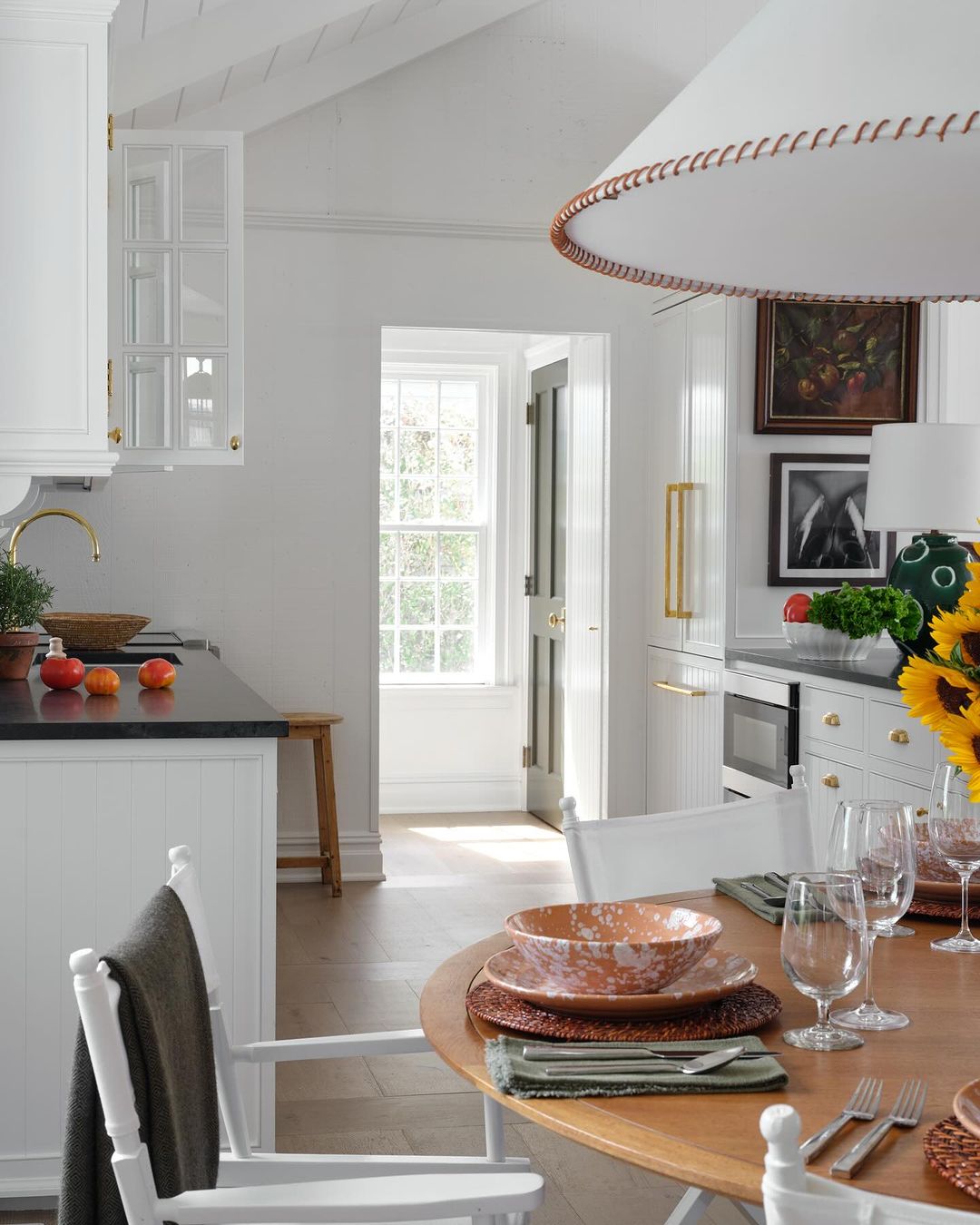 Well-lit home dining and kitchen area showcases a wood dining table, set dinnerware, natural light, and white kitchen cabinets.