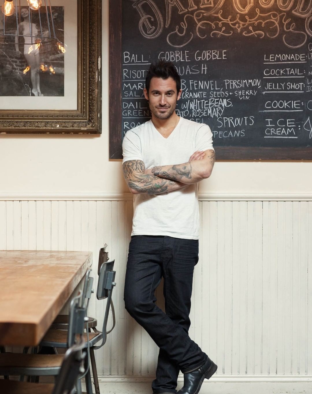 Adult male posing indoors with crossed arms in front of a wall-mounted chalkboard menu; natural light and medium camera angle.