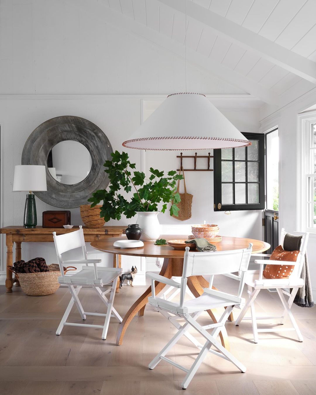 Dining room interior with round wooden table, white director's chairs, and decorative lighting. Natural light and neutral color palette.