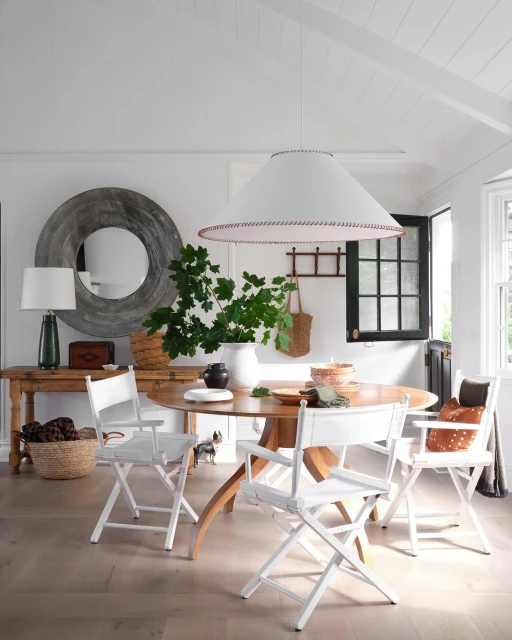 Dining room interior with round wooden table, white director's chairs, and decorative lighting. Natural light and neutral color palette.