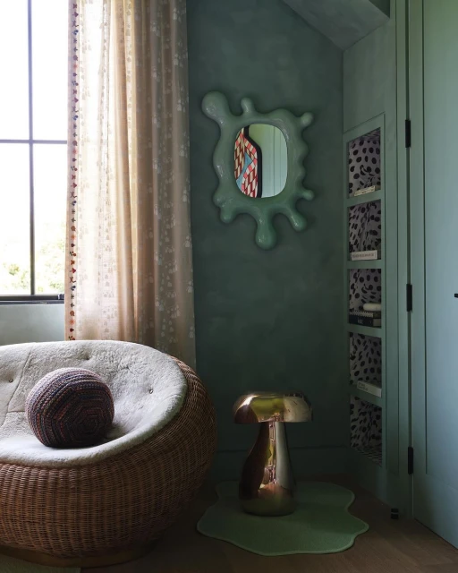 Interior view showcases a wicker chair with a brown spherical cushion, a shiny gold side table, and a light-green mirror on a green wall.