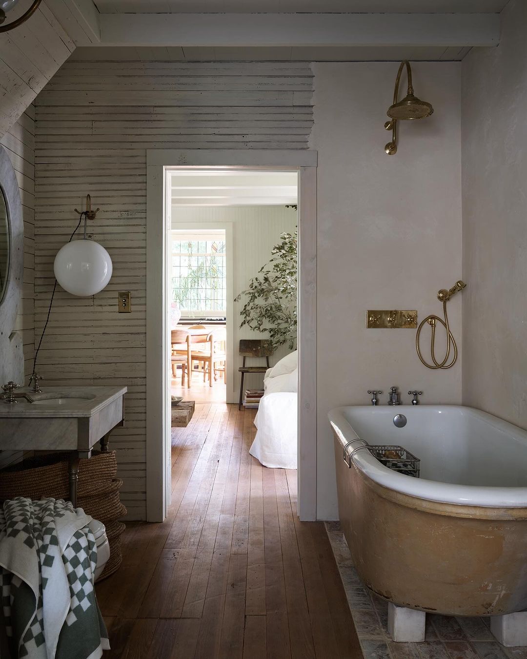 Rustic bathroom interior featuring a freestanding tub, vanity sink, and open doorway leading to a bedroom with a white bed and chairs.