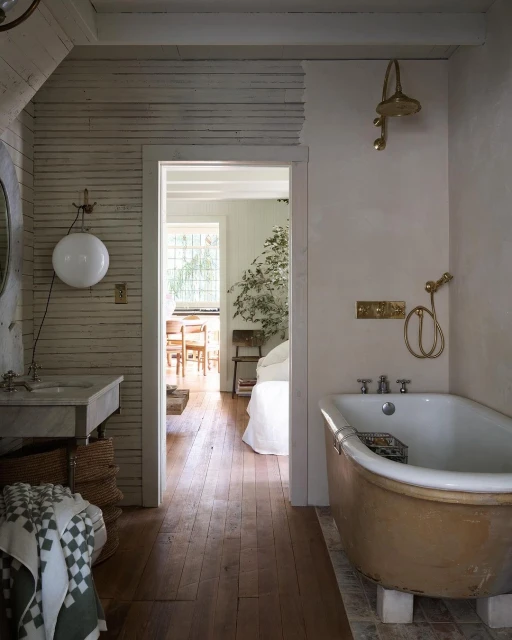 Rustic bathroom interior featuring a freestanding tub, vanity sink, and open doorway leading to a bedroom with a white bed and chairs.