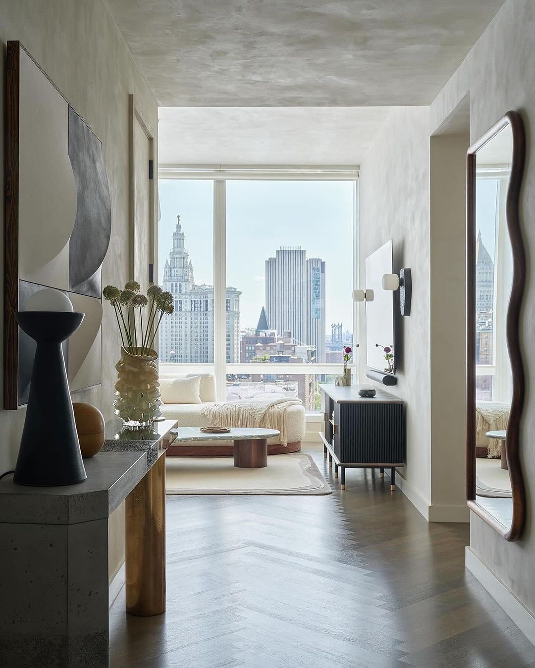 Living room interior with decor in neutral tones, city view through a large window, and chevron hardwood floors.