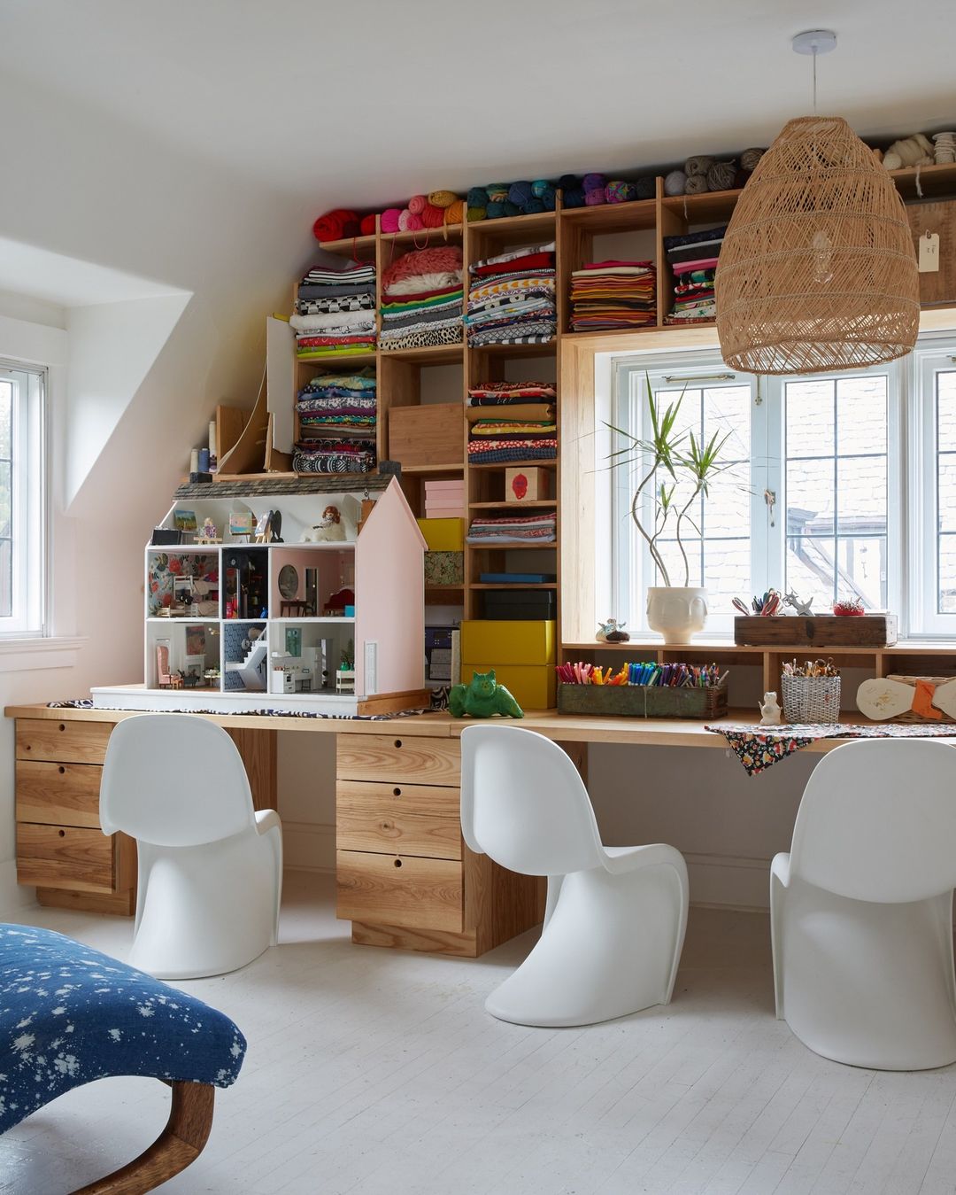 Craft room interior with a large wooden desk, white Eames chairs, shelving with fabric, yarn and craft supplies, and a pendant light.