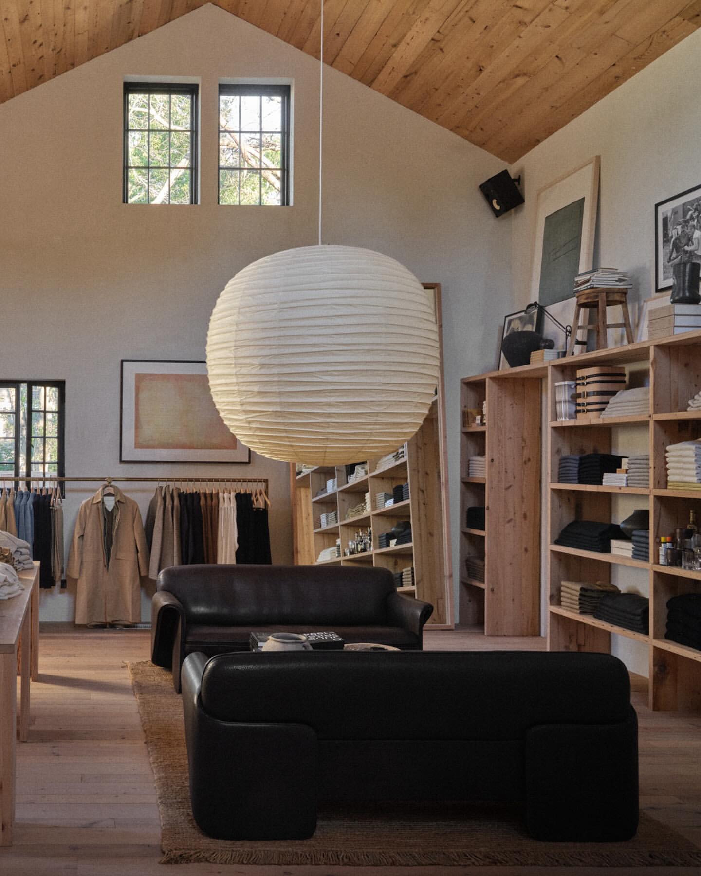 Interior of a stylish store with a paper lantern, clothes rack, wooden shelves, and dark leather furniture in a warmly lit space.