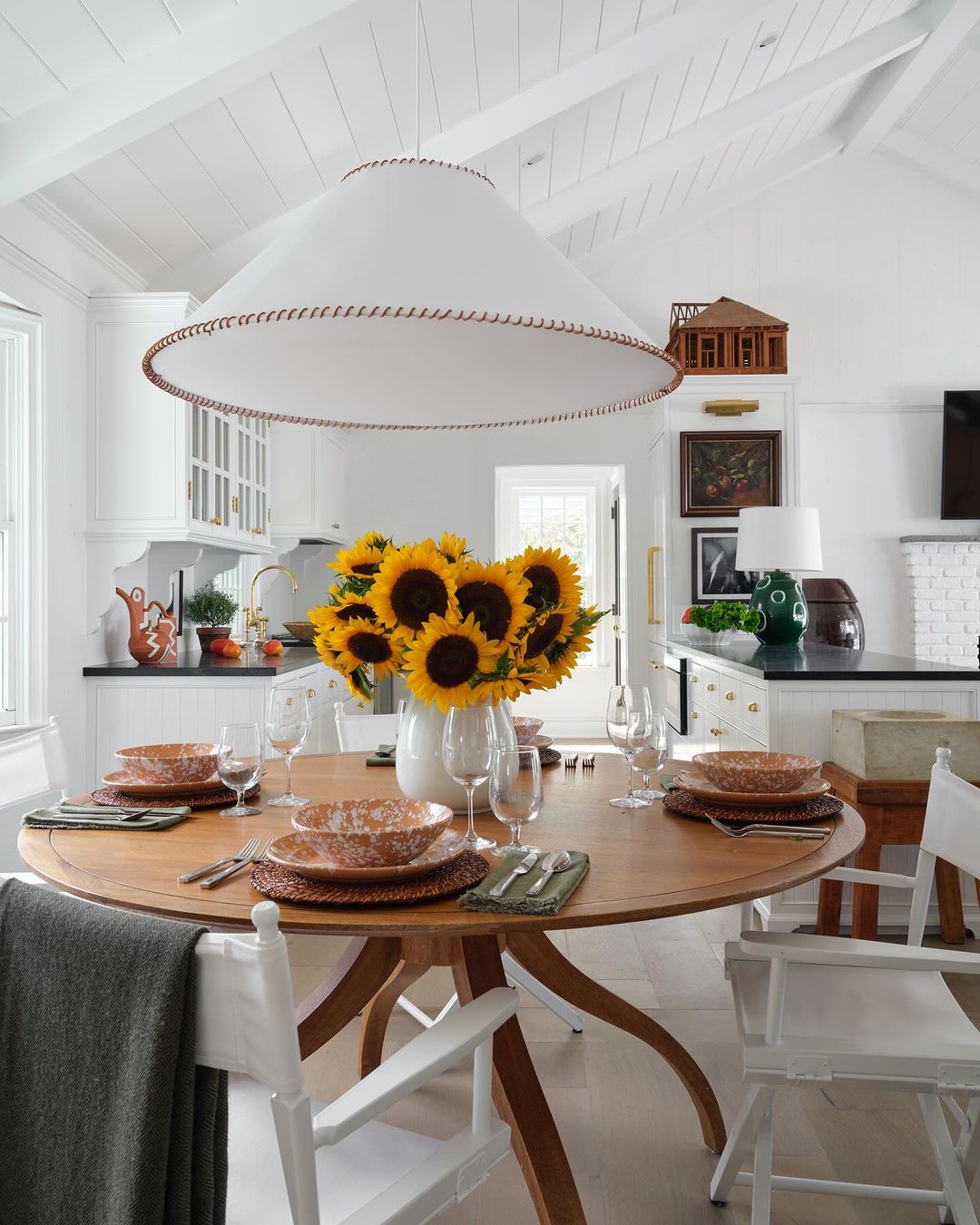 Dining room with a round wooden table set for a meal, featuring sunflowers in a white vase and a modern white pendant light fixture.