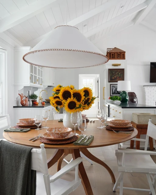 Dining room with a round wooden table set for a meal, featuring sunflowers in a white vase and a modern white pendant light fixture.