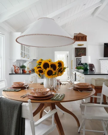 Dining room with a round wooden table set for a meal, featuring sunflowers in a white vase and a modern white pendant light fixture.