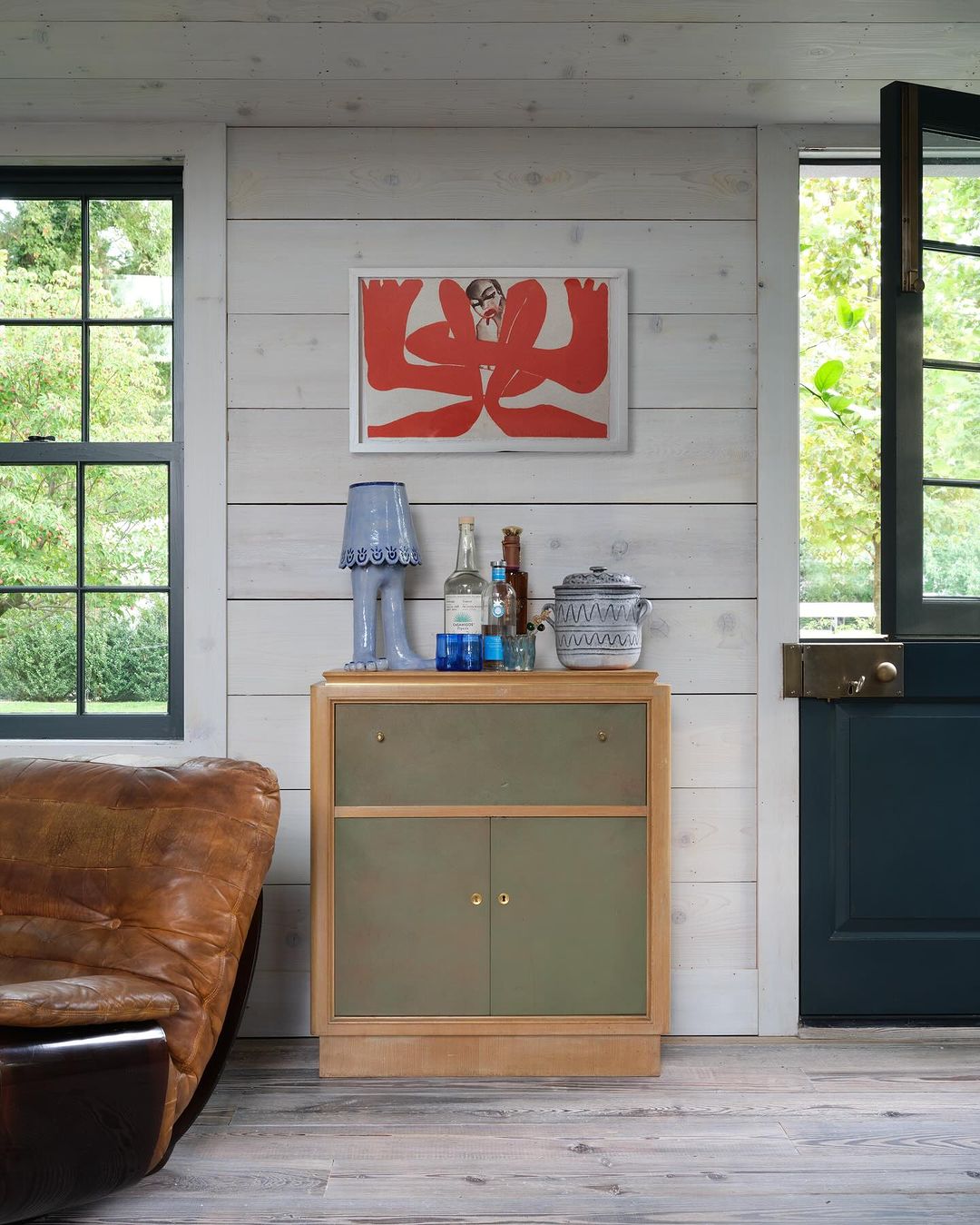 Indoor shot of a light wood cabinet with a table lamp, bottles, and a ceramic jar, set against a white shiplap wall with an abstract print.