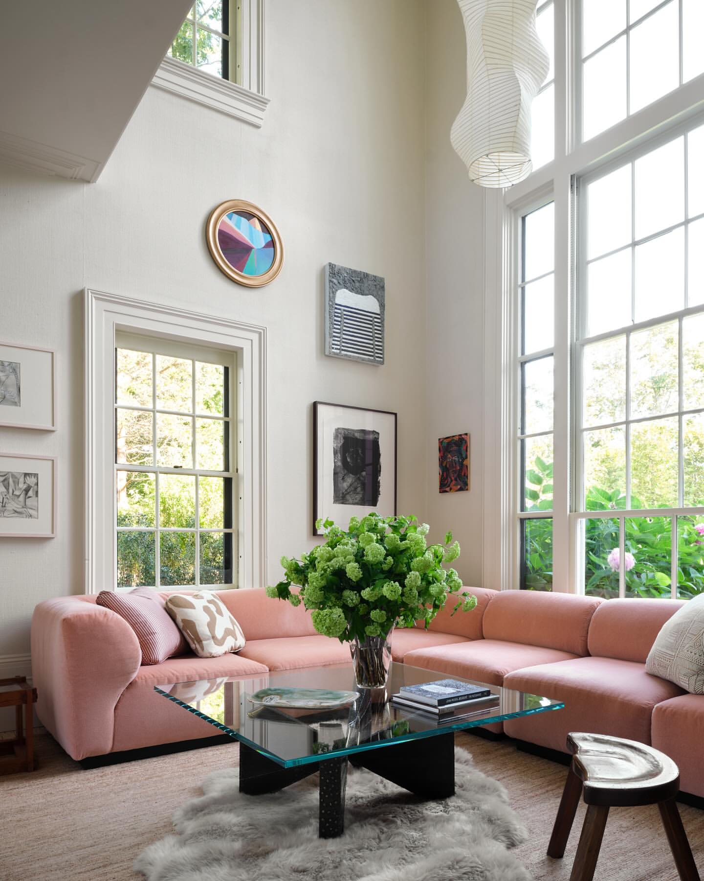 Living room interior with a pink sectional sofa, glass coffee table, and neutral decor, illuminated by natural light from large windows.