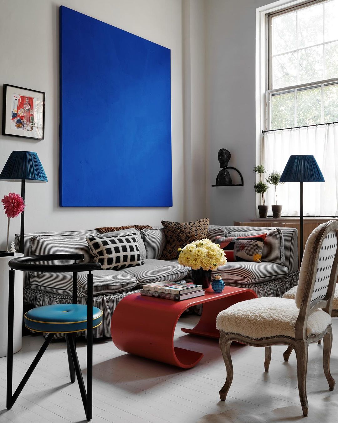 Living room interior with a blue painting above a grey couch, red table with flowers, a black side table and a patterned chair.