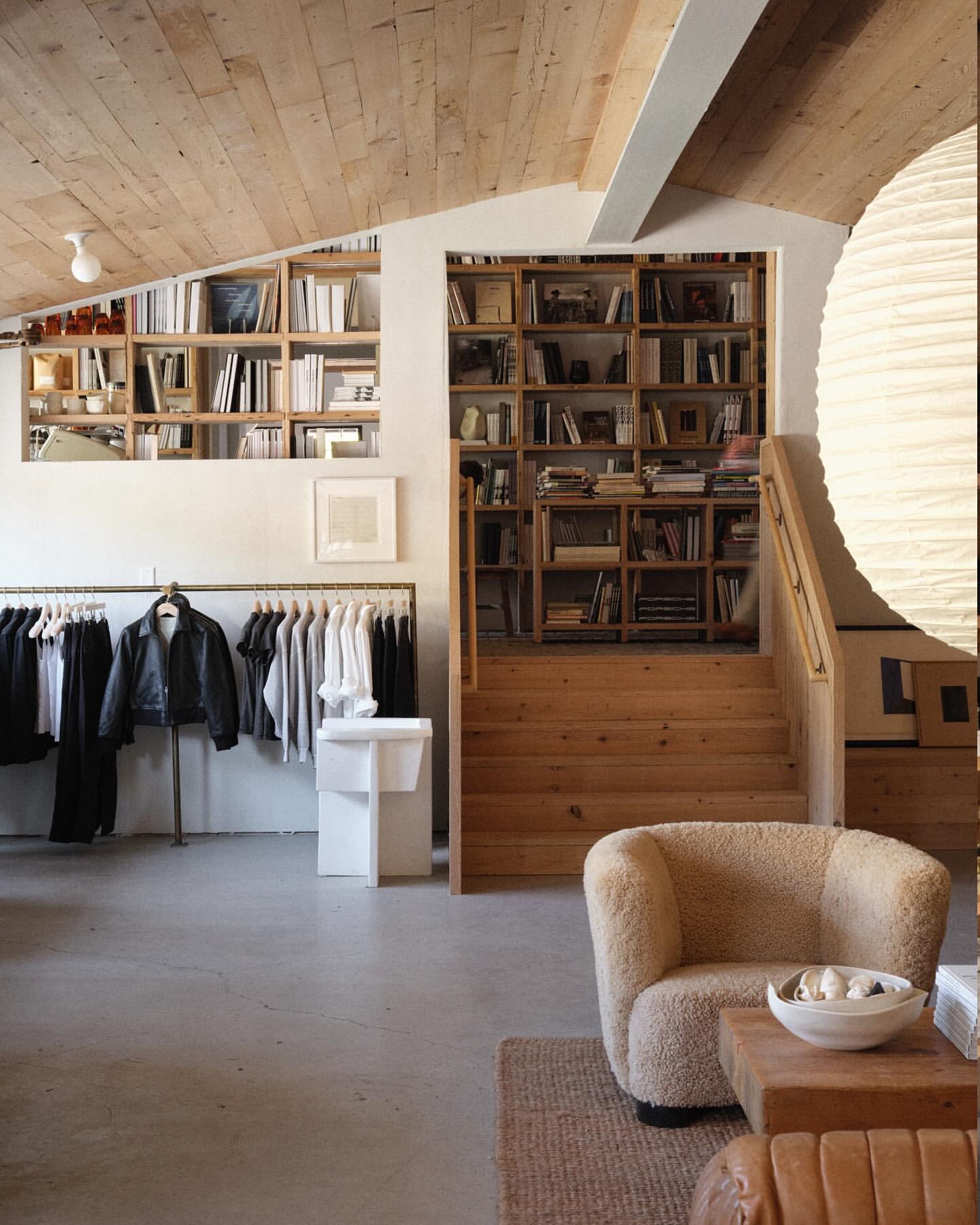Clothing store interior with shirts, jacket, and pants on rack and bookshelves on the back wall, in neutral tones and natural light.