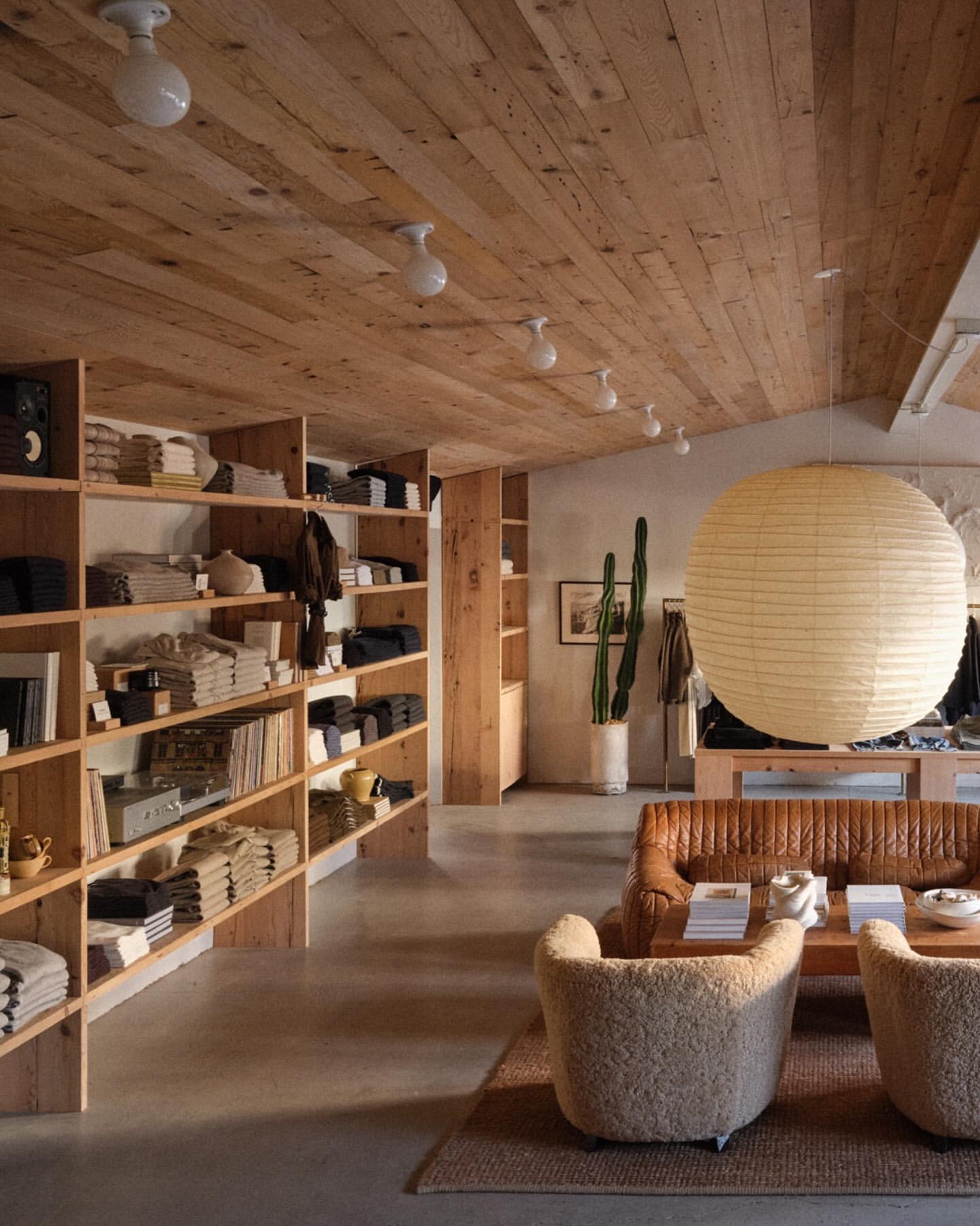 Interior of a retail store with shelves of folded garments, a tan leather sofa, two sherpa chairs, and a large paper lantern.