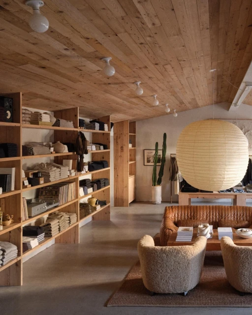 Interior of a retail store with shelves of folded garments, a tan leather sofa, two sherpa chairs, and a large paper lantern.