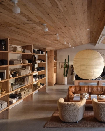 Interior of a retail store with shelves of folded garments, a tan leather sofa, two sherpa chairs, and a large paper lantern.