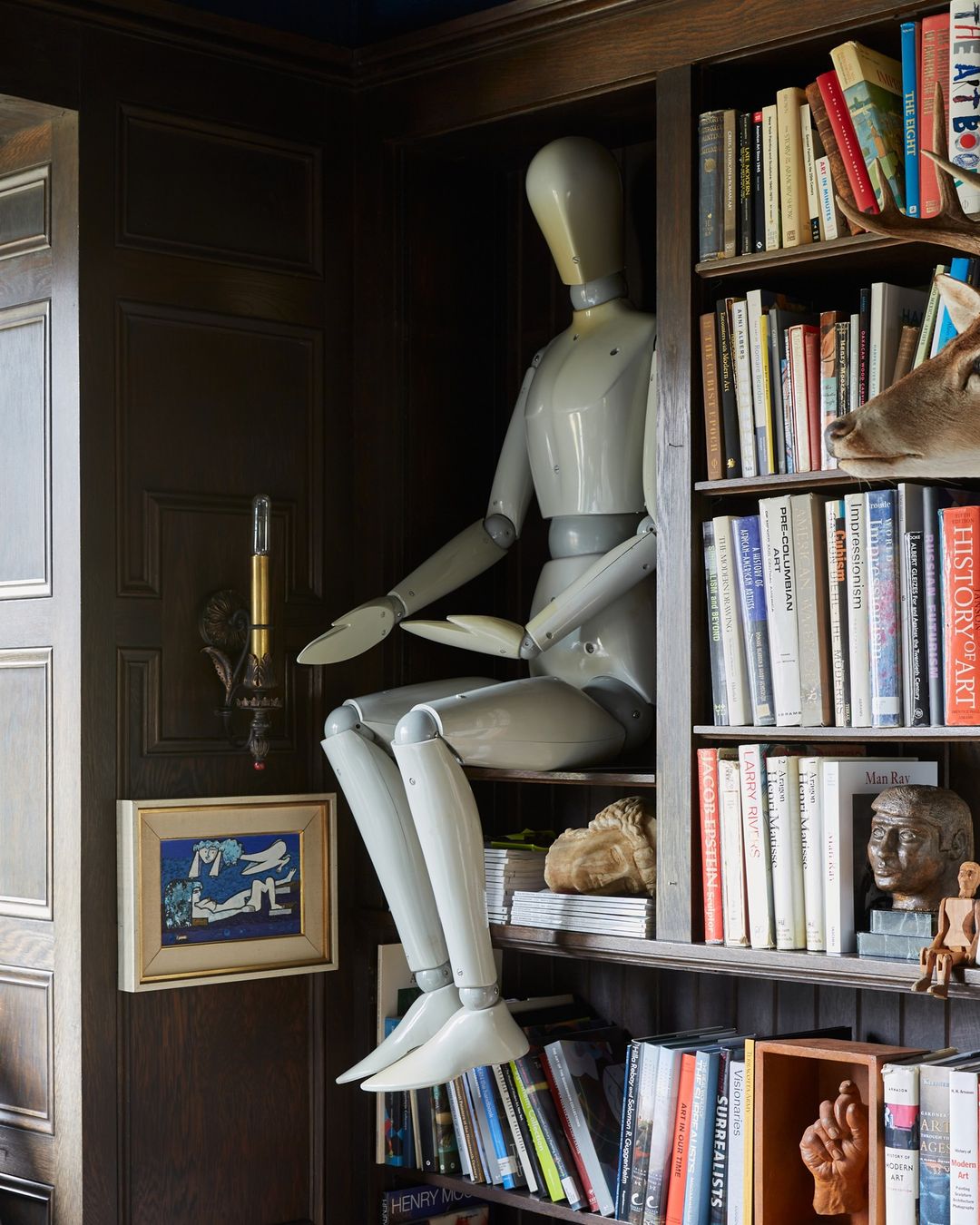 Wooden mannequin figure sits on a bookshelf filled with books and a deer head mount in a traditionally styled home interior.