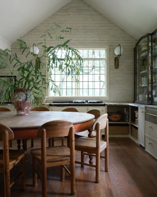 Warm-toned interior featuring a rustic round dining table surrounded by wooden chairs, accented by a large potted plant near a window.
