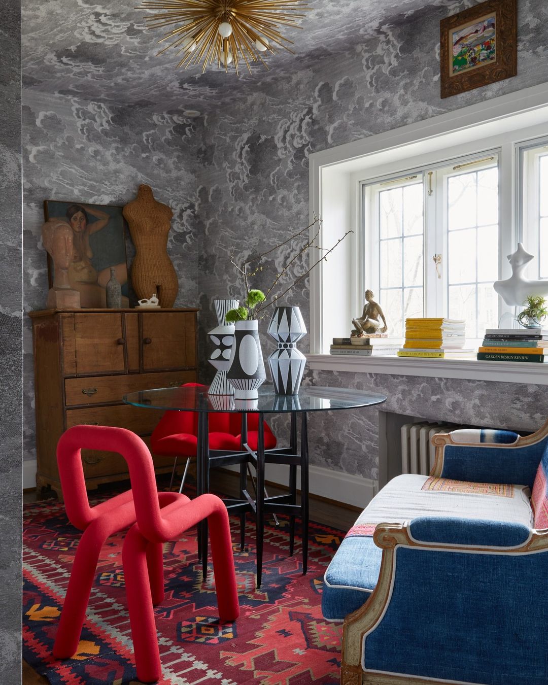 Interior view of a dining area with a red chair, table setting, artwork, patterned wallpaper, and an ornamental ceiling light fixture.