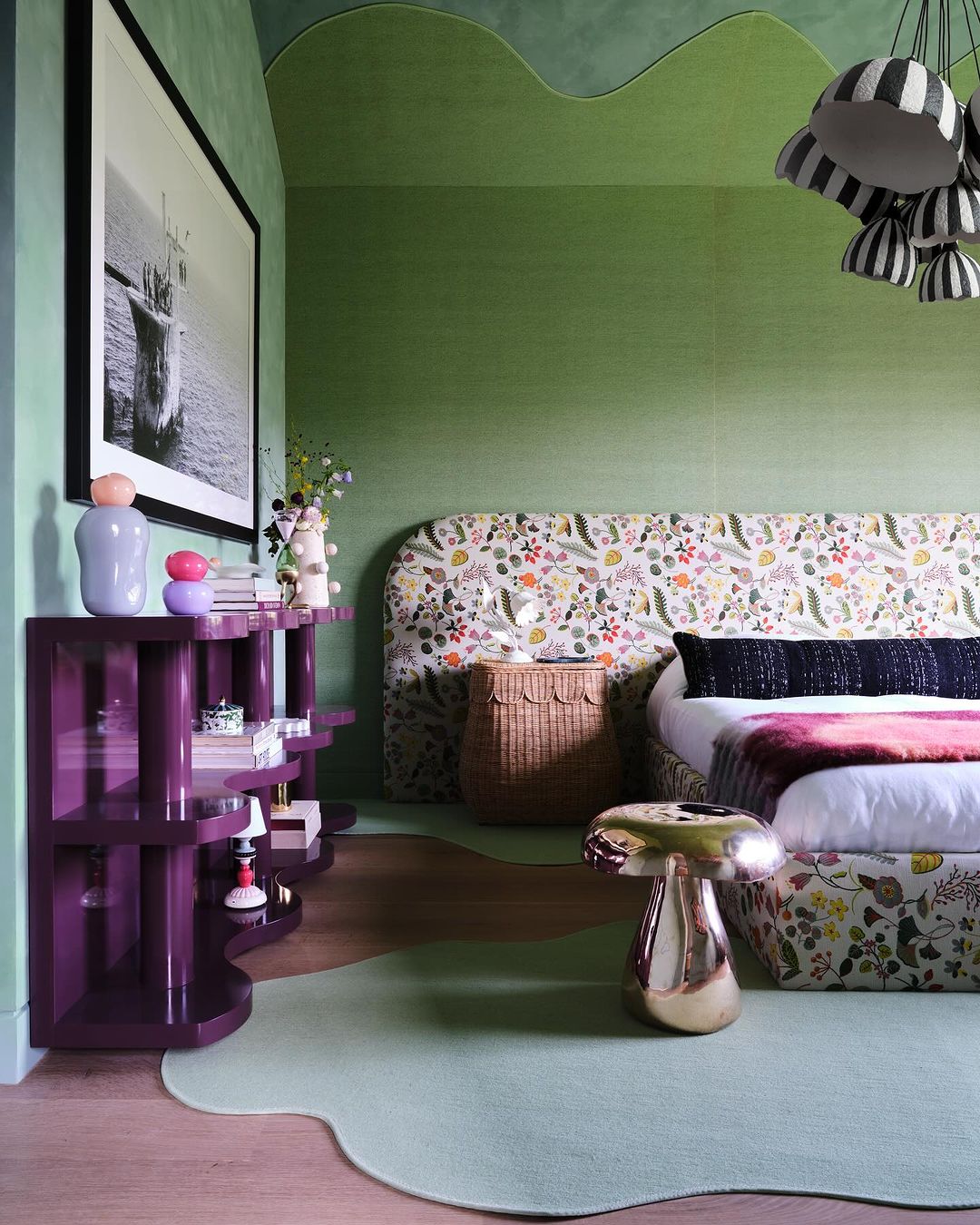 Bedroom interior featuring a bed with floral bedding, a purple console table, a mushroom stool, and green textured walls and trim.