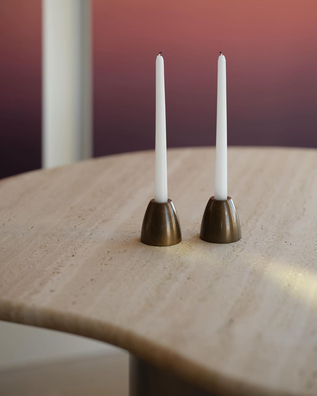 Two white taper candles in brass holders sit atop a round, beige table against a blurred gradient background of pinks and browns.