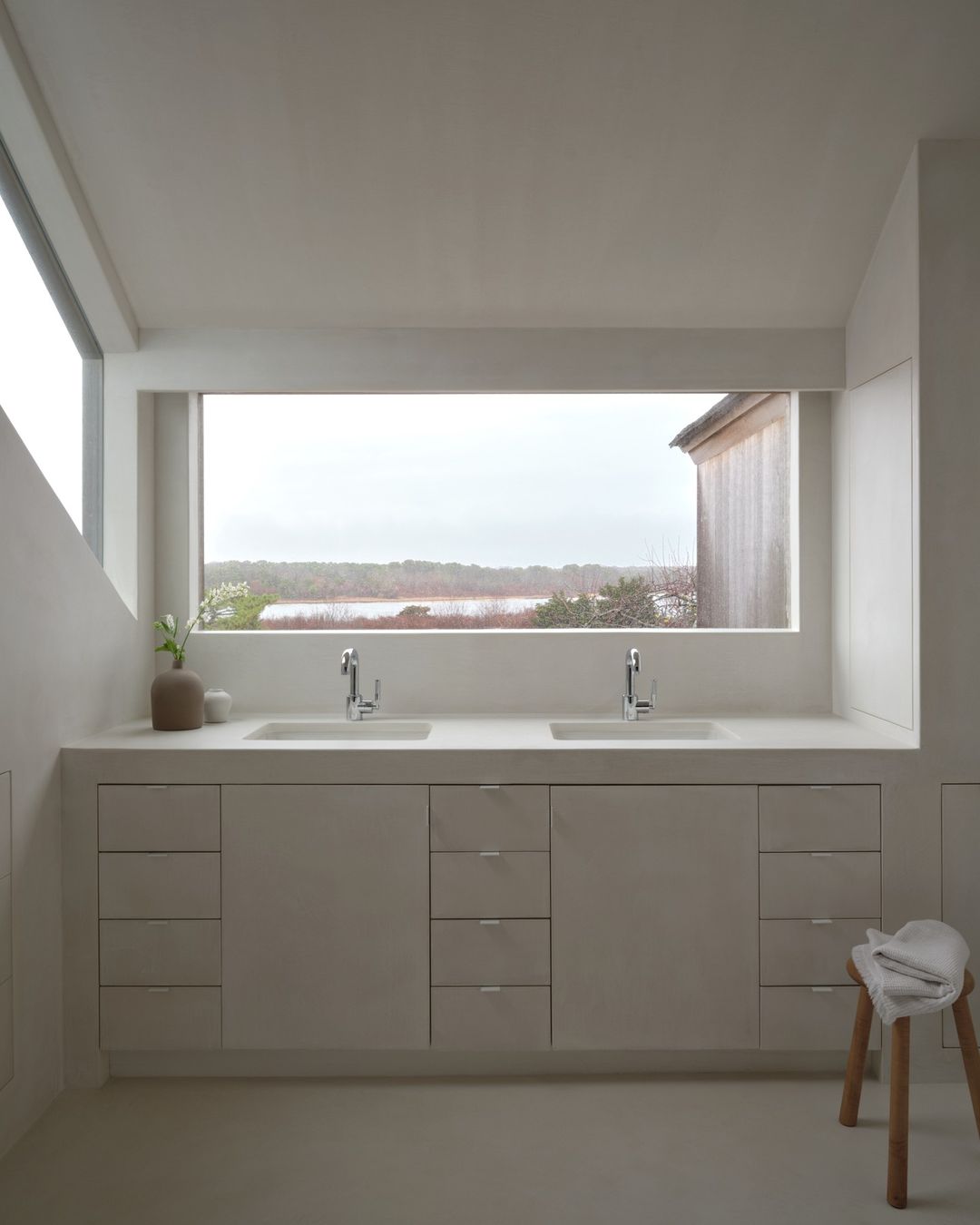 Bathroom interior with a double sink vanity, light color palette, large picture window overlooking a lake, and a wooden stool.