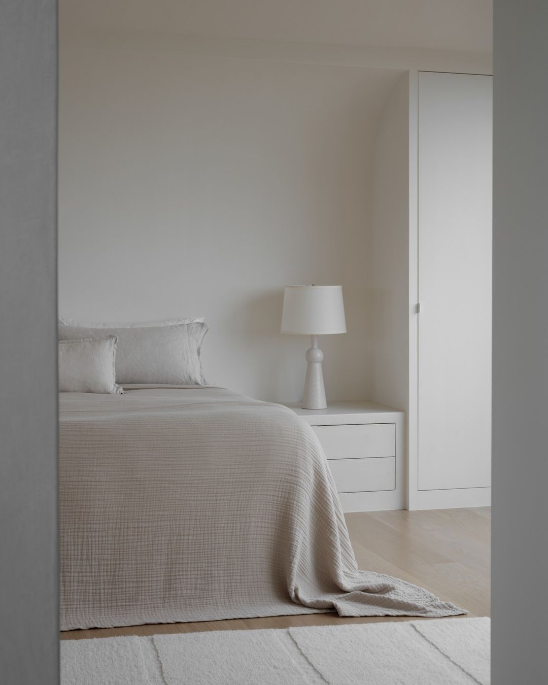 Minimalist bedroom with a neutral color palette, featuring a bed with white linens, a nightstand with a lamp, and a white carpet.