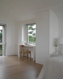 Minimalist modern bedroom interior with white walls, a wooden desk, a woven chair, and a tall built-in wardrobe.