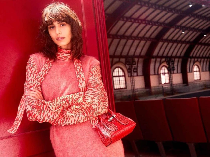 Fashionable woman in pink knitted sweater and dress with red purse poses in a red-themed train station with arched windows.