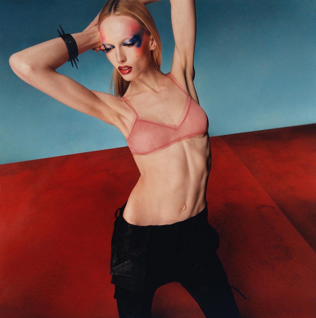 Fashion shot of a slim woman with blonde hair, wearing a pink lingerie top and black pants, striking a pose against a red and blue background.