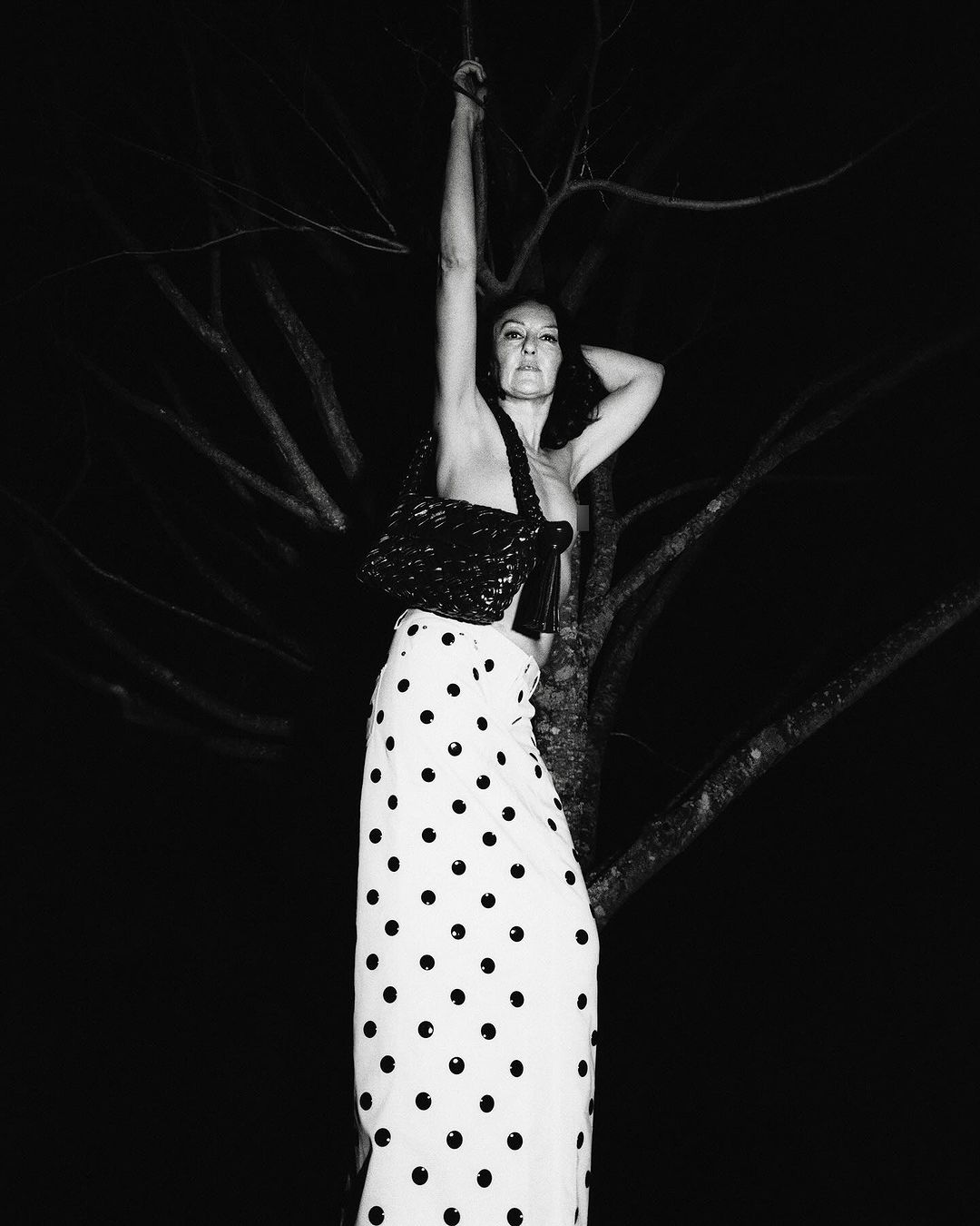 Woman in polka dot skirt holding a black handbag posing against a dark tree at night by Remi Lamande