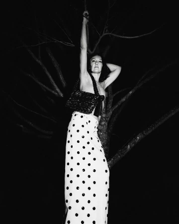 Woman in polka dot skirt holding a black handbag posing against a dark tree at night by Remi Lamande