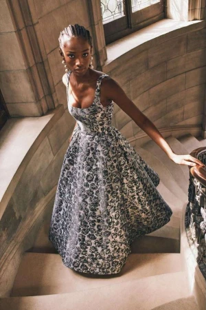 Black woman descending stone stairs wearing a patterned square-neck evening dress and pearl earrings, photographed by Rémi Lamandé.