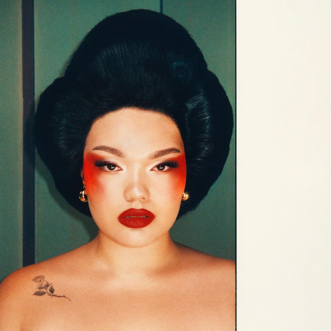 Portrait of an adult female with dramatic red and white makeup, gold earrings, and black updo hairstyle, in front of a green wall.