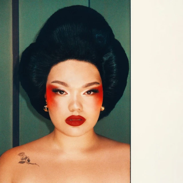 Portrait of an adult female with dramatic red and white makeup, gold earrings, and black updo hairstyle, in front of a green wall.