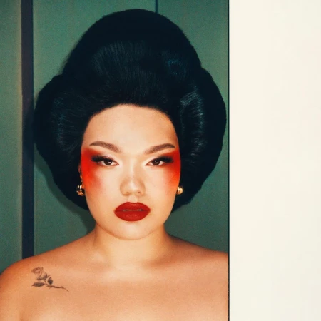 Portrait of an adult female with dramatic red and white makeup, gold earrings, and black updo hairstyle, in front of a green wall.