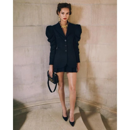Woman wearing structured black blazer with puffed shoulders and a handbag standing against stone wall