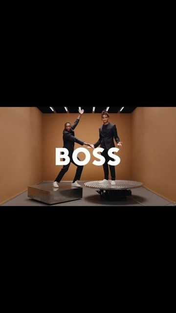 Two male fashion models in suits pose in a brightly lit studio, standing on platforms beneath the large white 'BOSS' logo.