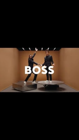 Two male fashion models in suits pose in a brightly lit studio, standing on platforms beneath the large white 'BOSS' logo.