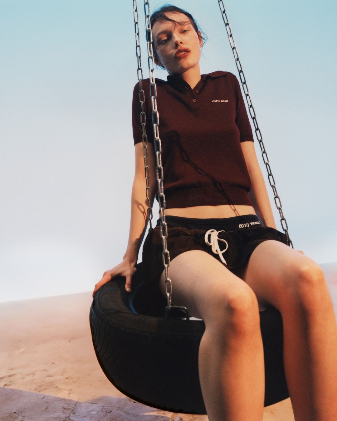 A young woman sits on a tire swing in an outdoor desert-like setting, wearing a burgundy polo shirt and black shorts with branding details.