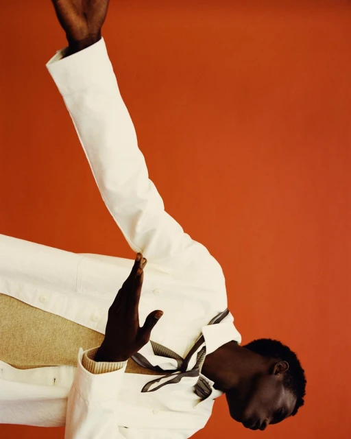 Portrait of a dark-skinned adult male in a cream-colored shirt, standing against a muted red background in a fashion editorial style.