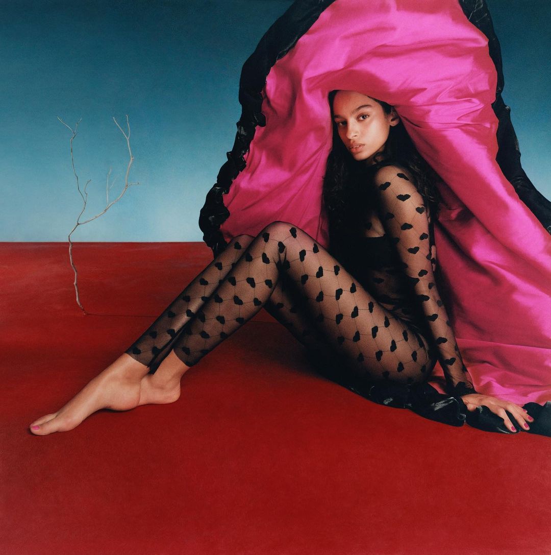 Elegant woman in sheer patterned jumpsuit seated on a red surface with a magenta and black fabric draped behind her against a blue wall.