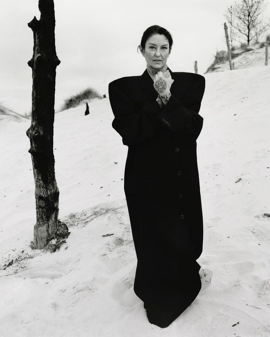 Portrait of a mature woman with tattoos wearing a black coat in a sandy, natural setting with sparse vegetation and a grey sky.