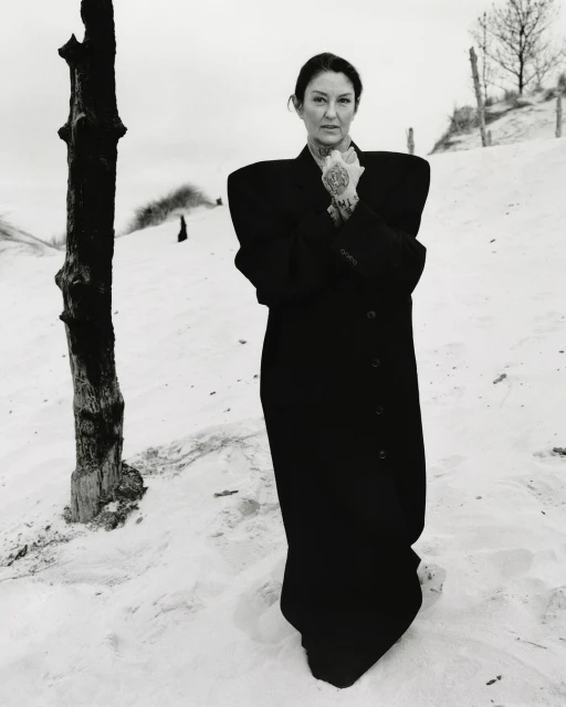 Portrait of a mature woman with tattoos wearing a black coat in a sandy, natural setting with sparse vegetation and a grey sky.