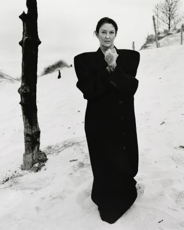 Portrait of a mature woman with tattoos wearing a black coat in a sandy, natural setting with sparse vegetation and a grey sky.