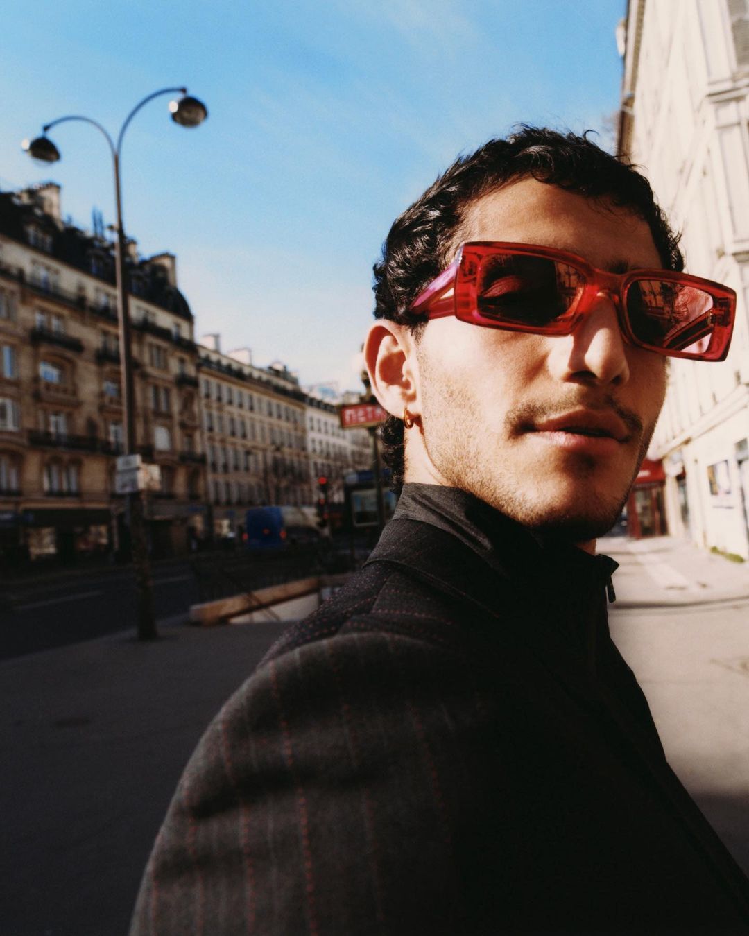 Portrait of a stylish man wearing red sunglasses on a city street with buildings and streetlights under a clear blue sky.