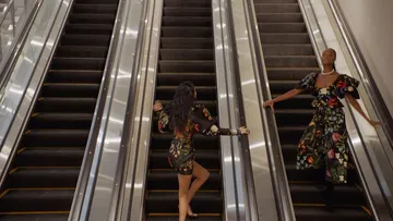 Two women in floral-patterned outfits on escalators, ascending in an indoor setting, with bright lighting enhancing the fabric details.