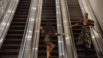 Two women in floral-patterned outfits on escalators, ascending in an indoor setting, with bright lighting enhancing the fabric details.