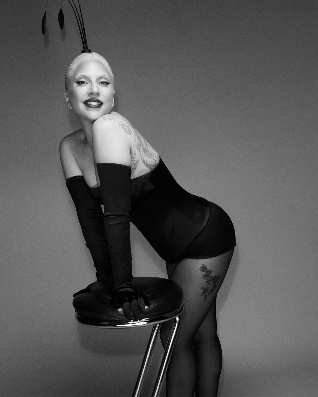 Black and white portrait of an adult woman with platinum blonde hair, leaning on a chrome stool with feather headpiece and tattoos.